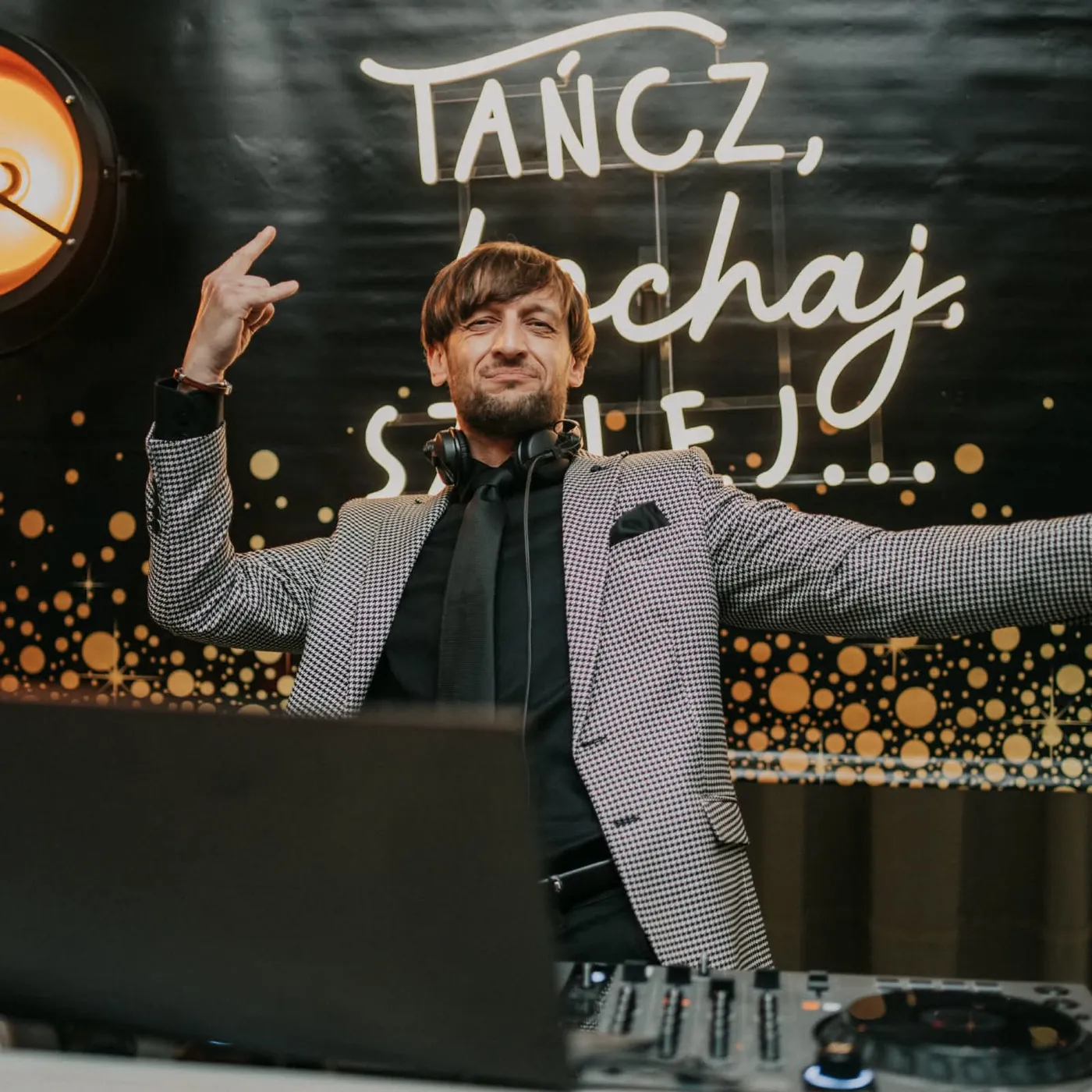 DJ Bartosz Zdunek przy konsoli na tle neonu „Tańcz, kochaj, szalej”