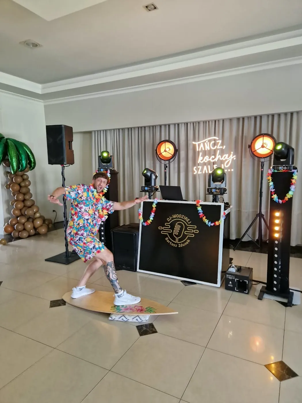 Wodzirej w hawajskiej koszuli „surfuje” na desce przed stanowiskiem DJ-a