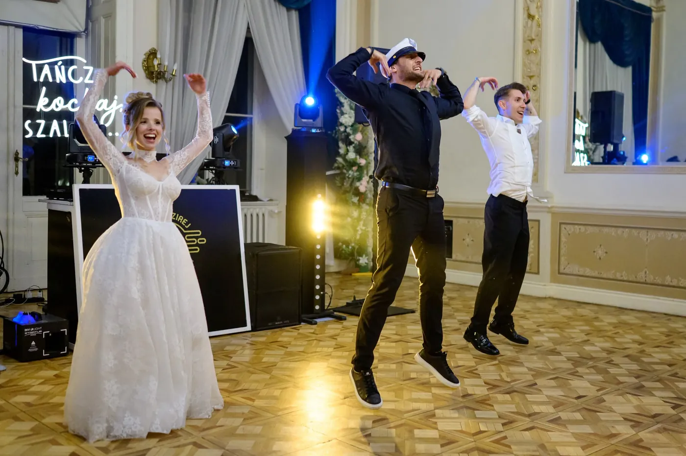Panna młoda, wodzirej w marynarskiej czapce i gość tańczą w pałacowej sali pod neonem